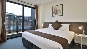 A hotel room with a large bed, a nightstand with a lamp, a framed artwork on the wall, and a view of the outside through a window with curtains. - Quality Apartments Dandenong (Dandenong)