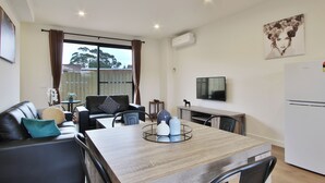 A modern living room with a dining area, a flat-screen TV mounted on the wall, and a large window with curtains. - Quality Apartments Dandenong (Dandenong)