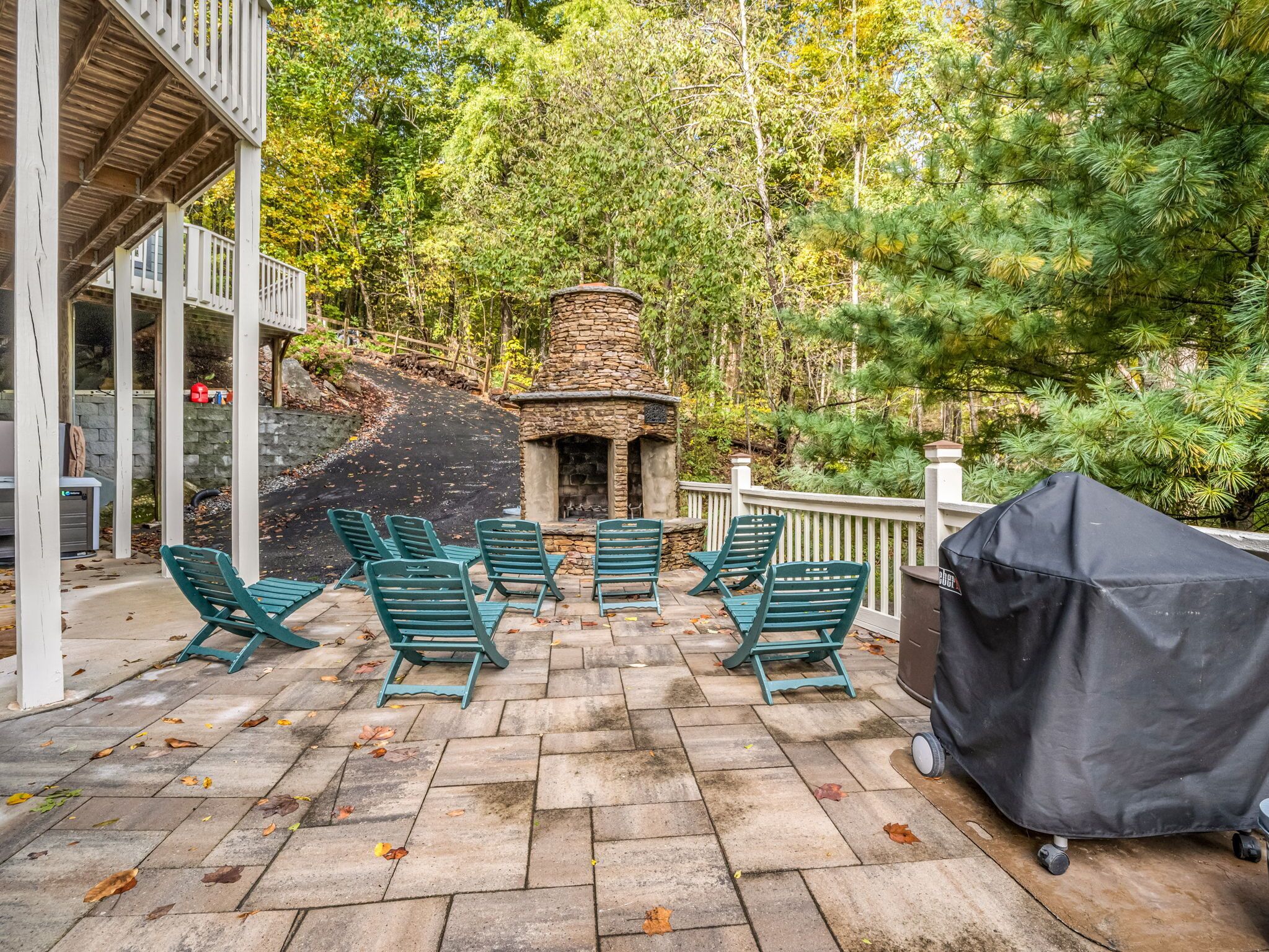 Une terrasse dallée de pierres, avec des chaises vertes et une cheminée en pierre, le tout entouré d’arbres.