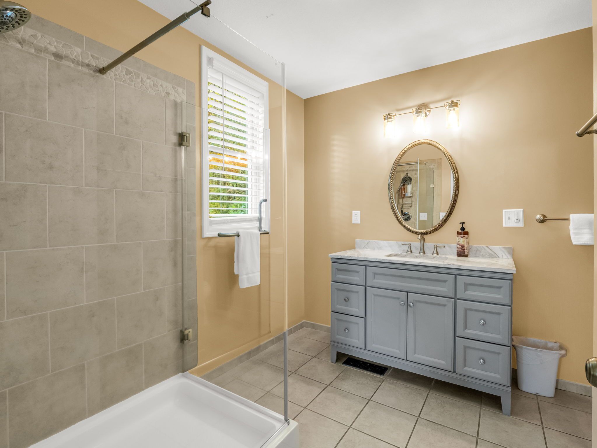 Une salle de bain avec une douche, un meuble-lavabo avec un miroir et une fenêtre avec des stores.