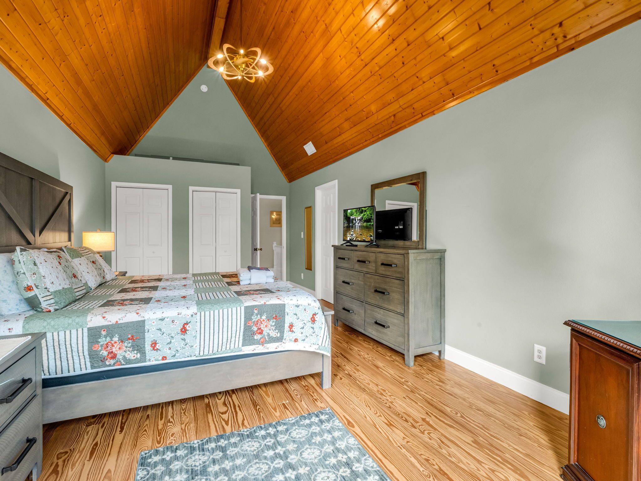 A bedroom with a wooden ceiling, a bed with a floral quilt, a dresser, and a television on top.