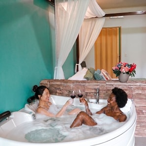 A room with a bathtub, a mirror, a potted plant, and a bed with pillows and a blanket. - Yellow Tree Suites (La Fortuna)