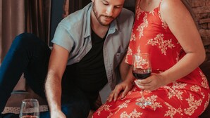 Un couple est assis sur un lit, une boîte de pizza et un verre à vin sont posés sur le sol.