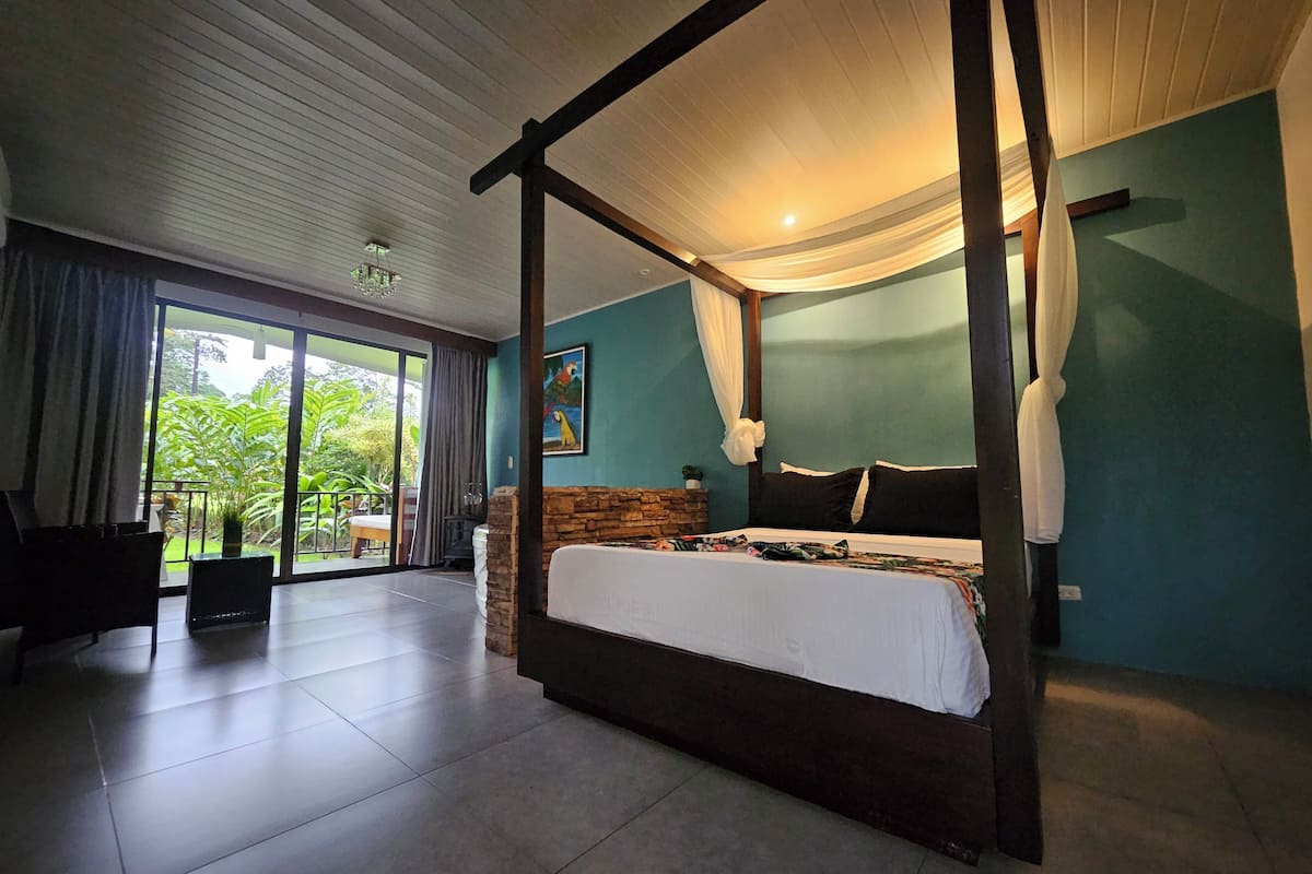 A four-poster bed with a stone accent wall, a large window with curtains, and a view of greenery outside.