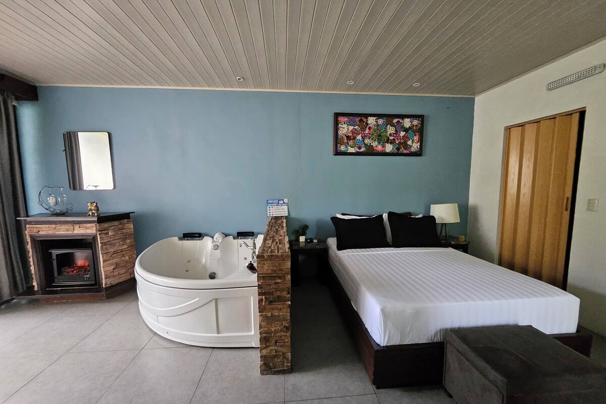 A bedroom with a bed, a nightstand, a lamp, a painting, a fireplace, and a bathtub.
