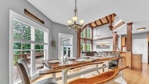 Un amplio comedor con una gran mesa de madera y bancos, una araña y ventanas grandes con persianas.