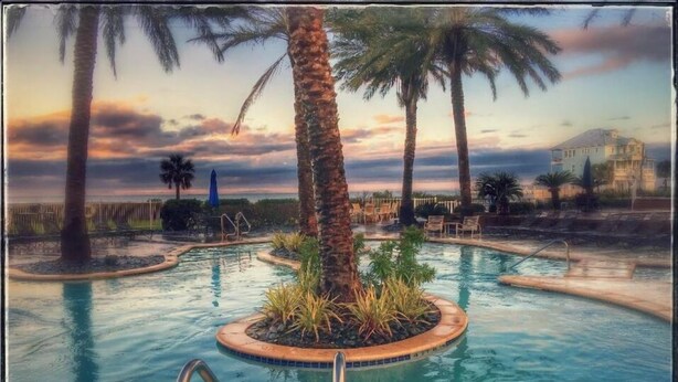 A swimming pool with palm trees and lounge chairs, surrounded by a fence and a building in the background. - Newly renovated Bayfront Condo, Beach Club, Pools, Hot Tubs, Sunsets and More!! (Galveston)