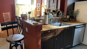 Een compacte keuken met een houten aanrecht, een RVS spoelbak en een ingebouwde vaatwasser. In de eethoek staan een barkruk en een stoel.