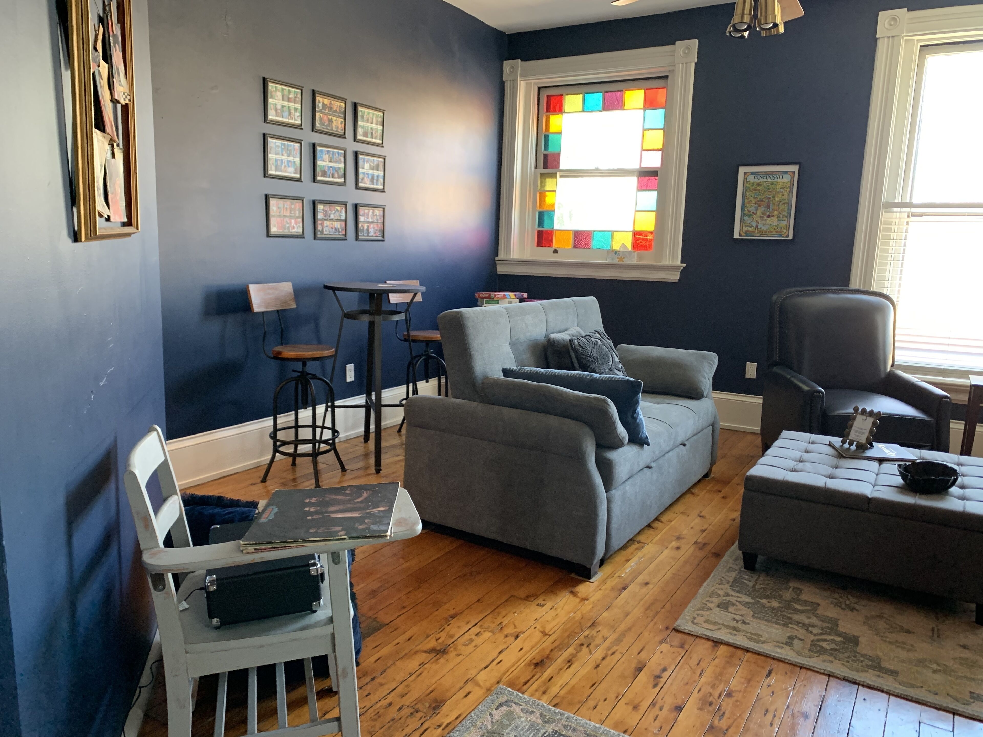 A living room with a grey sofa, a small table, two bar stools, a white chair, and a large window with a stained glass design.