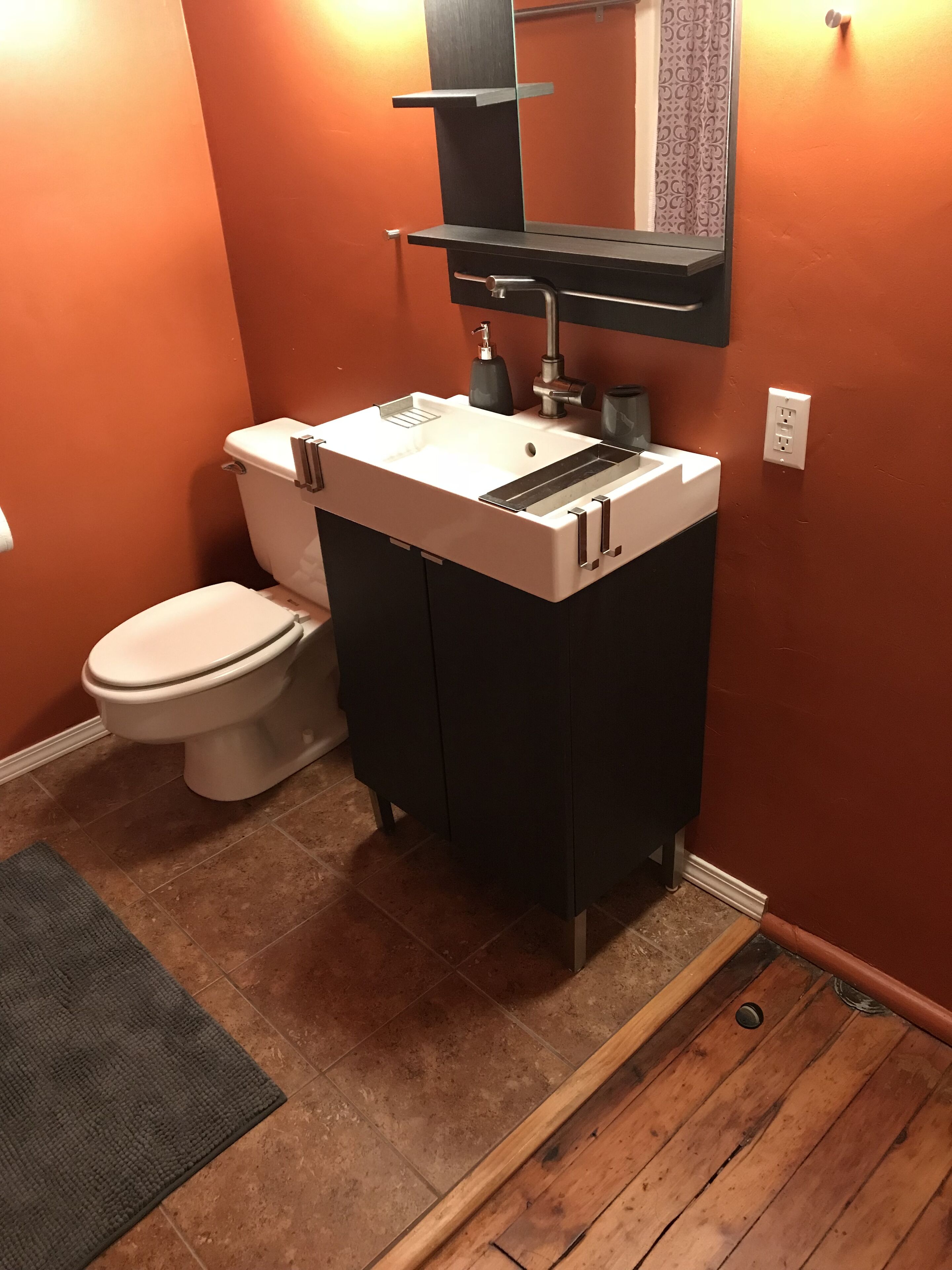 Une salle de bain avec des toilettes blanches, un lavabo noir avec un miroir au-dessus, et un plancher en bois.