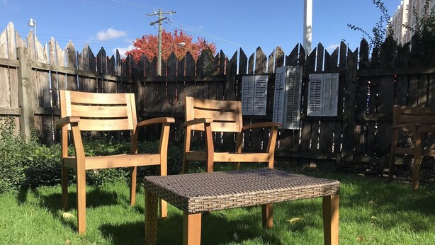 Twee houten stoelen en een geweven tafel in een gazon, met een houten hek op de achtergrond.