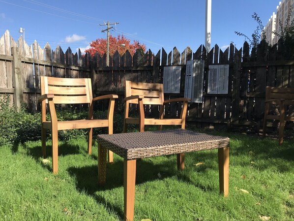 Two wooden chairs and a woven table in a grassy outdoor area with a wooden fence in the background. - Tres House - an experience, three floors high. (Covington)