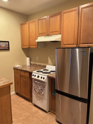 A kitchen with wooden cabinets, a stainless steel refrigerator, a stove, and a tiled floor. - Private Cottage (Mastic Beach)