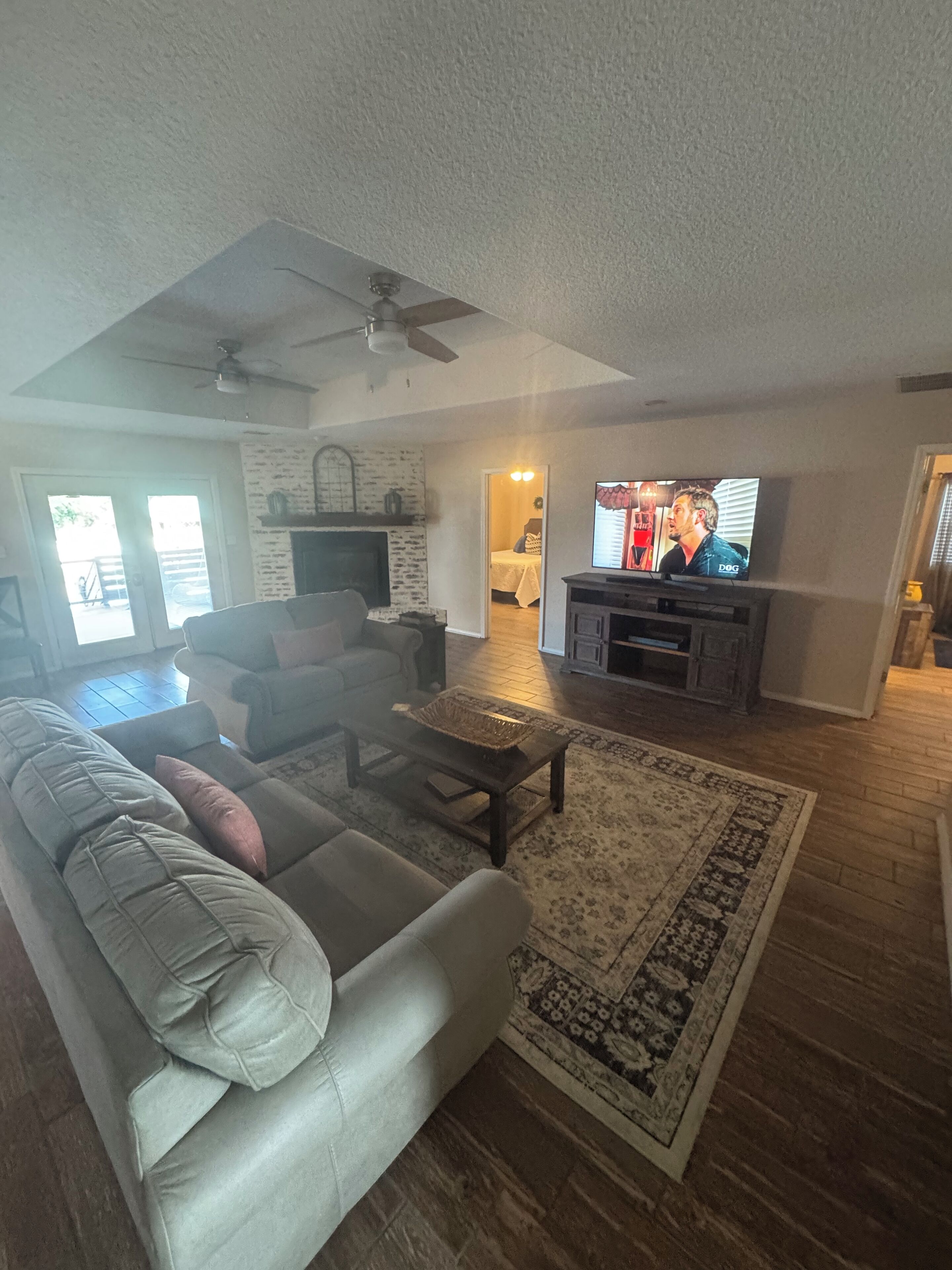 A living room with a sofa, coffee table, and TV. There is a fireplace, a ceiling fan, and a rug.