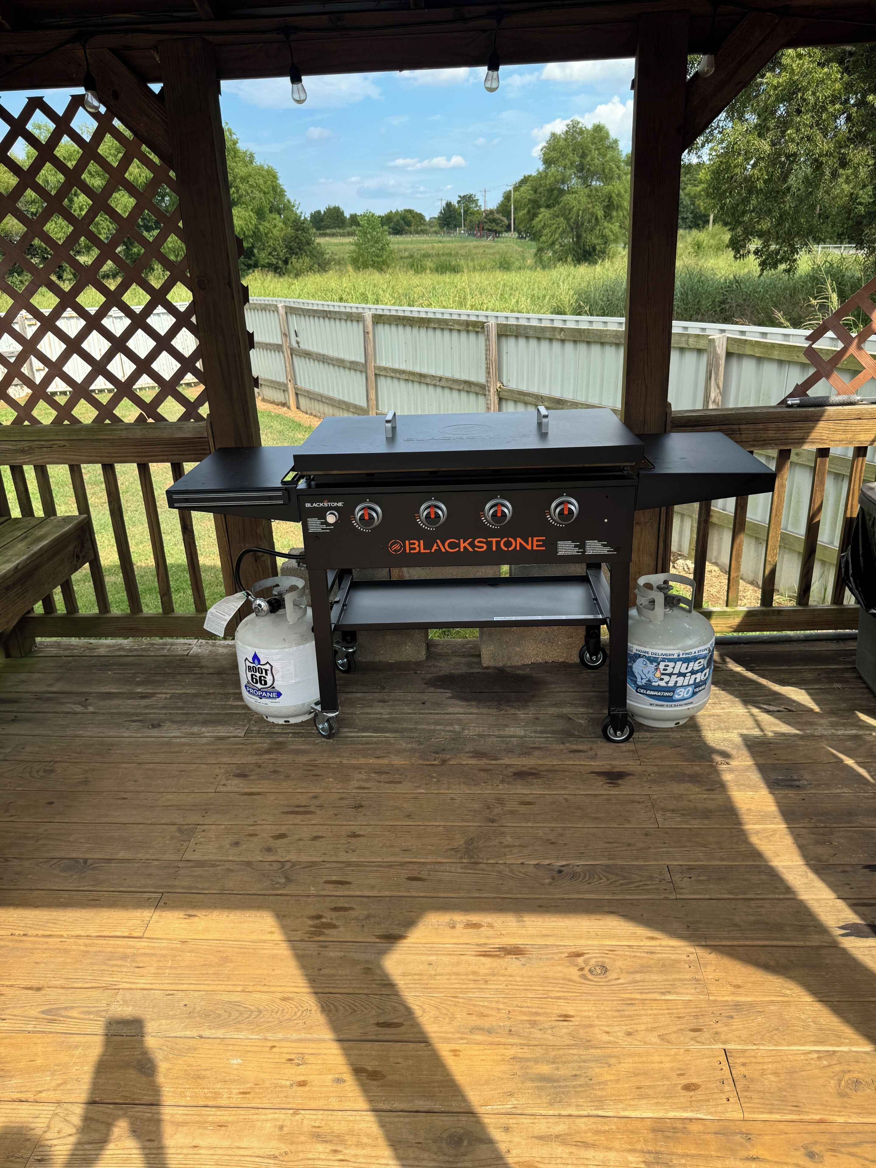 Una griglia da barbecue esterna con bombole di propano, posizionata su un patio in legno con vista su un prato.