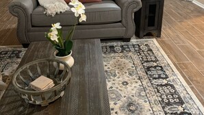 Een woonkamer met een grijze bank, een bijzettafeltje met een bloevas, een kamerbreed tapijt en een open haard met decoratieve items.