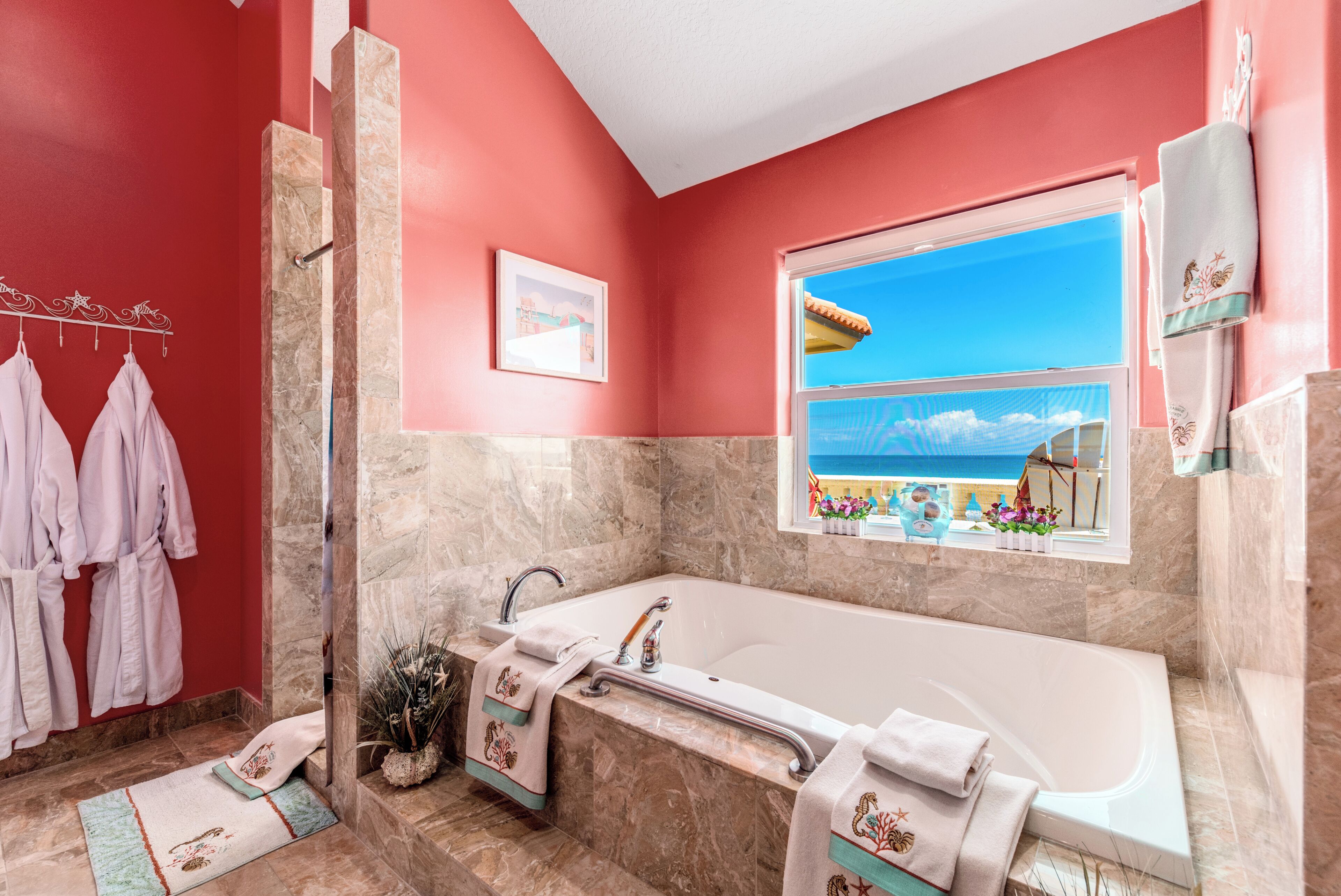 Un baño con bañera, ducha, ventana con vistas a la playa y toallas en un escurreplatos.