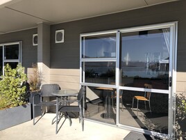A patio with a table and chairs, a large sliding glass door, and a view of a body of water and mountains.