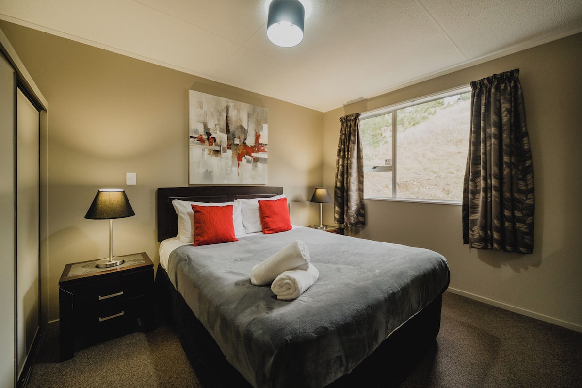 A hotel room with a bed, two bedside lamps, a window with curtains, a painting on the wall, and a small table with a lamp.