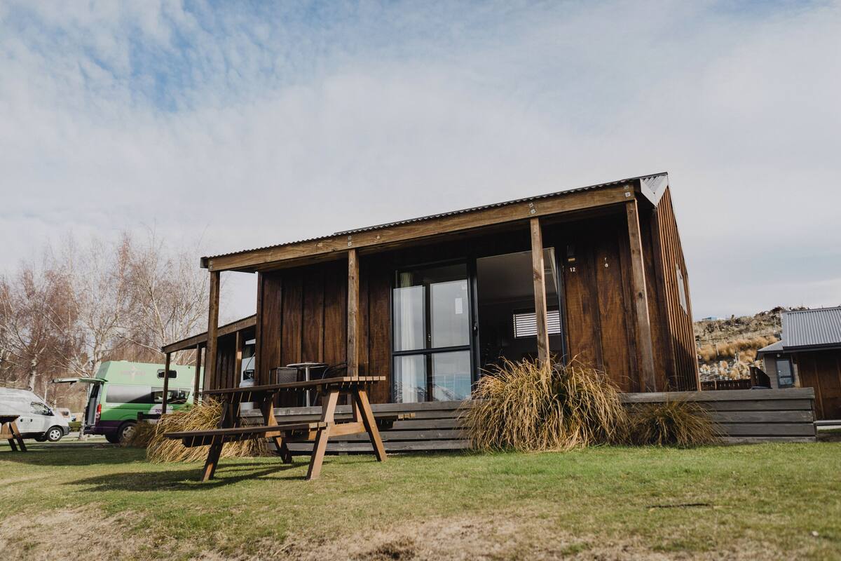 A wooden cabin with a large window, a picnic table, and a truck parked outside.