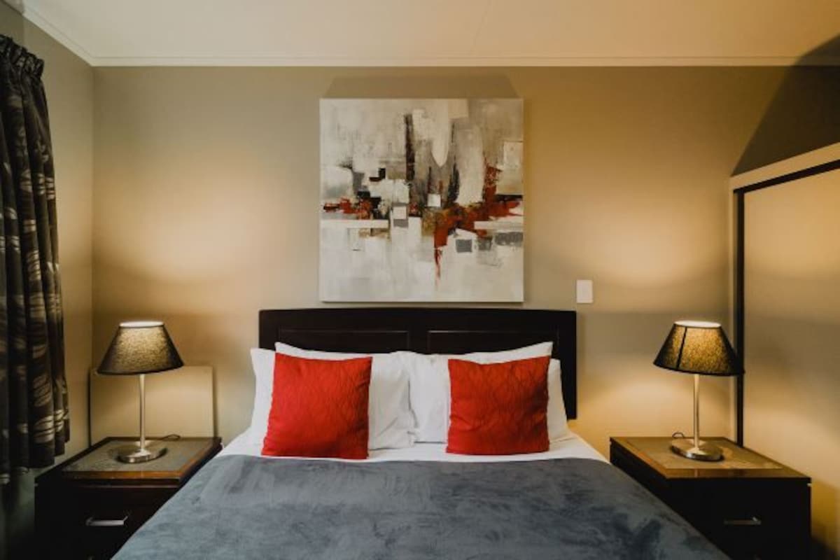 A hotel room with a bed, two bedside tables with lamps, a large abstract painting on the wall, and a window with curtains.
