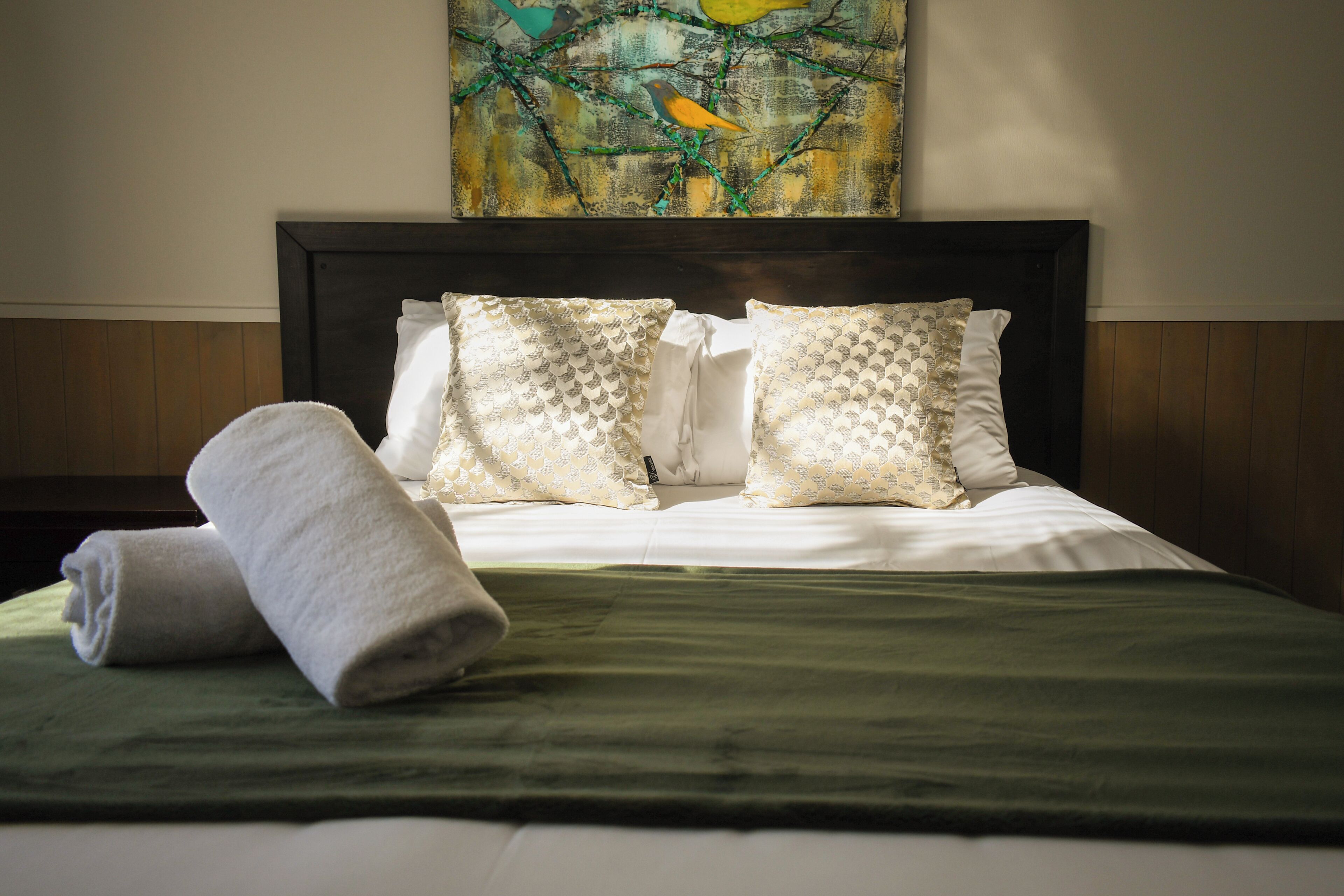 A neatly made bed with a green bedspread and two patterned pillows, a rolled white towel, and a painting above the bed.