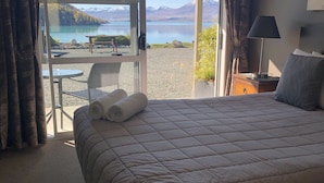 A hotel room with a large bed, a desk with a lamp, and a view of a lake and mountains through a glass door.