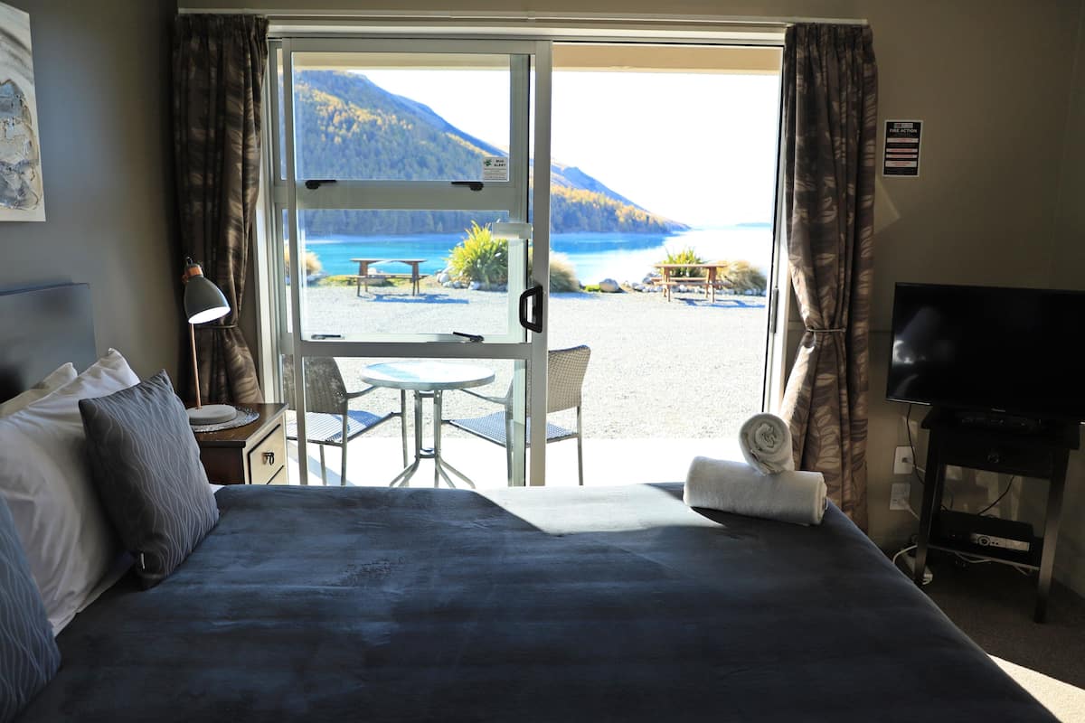 A hotel room with a bed, a TV, a desk with a lamp, and a view of a beach and mountains.