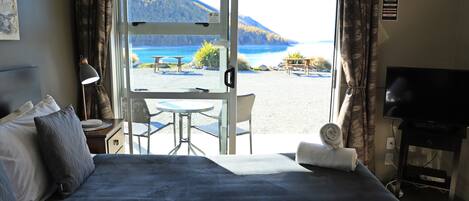 A hotel room with a bed, a TV, a desk with a lamp, and a view of a beach and mountains.
