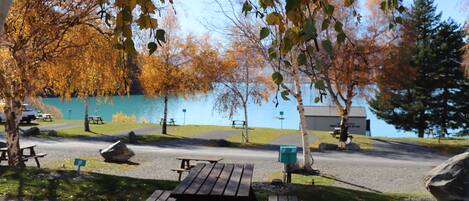 Ein Picknickbereich mit Bänken und Tisch, umgeben von Bäumen mit Herbstlaub, in der Nähe von Wasser und einem Gebäude.