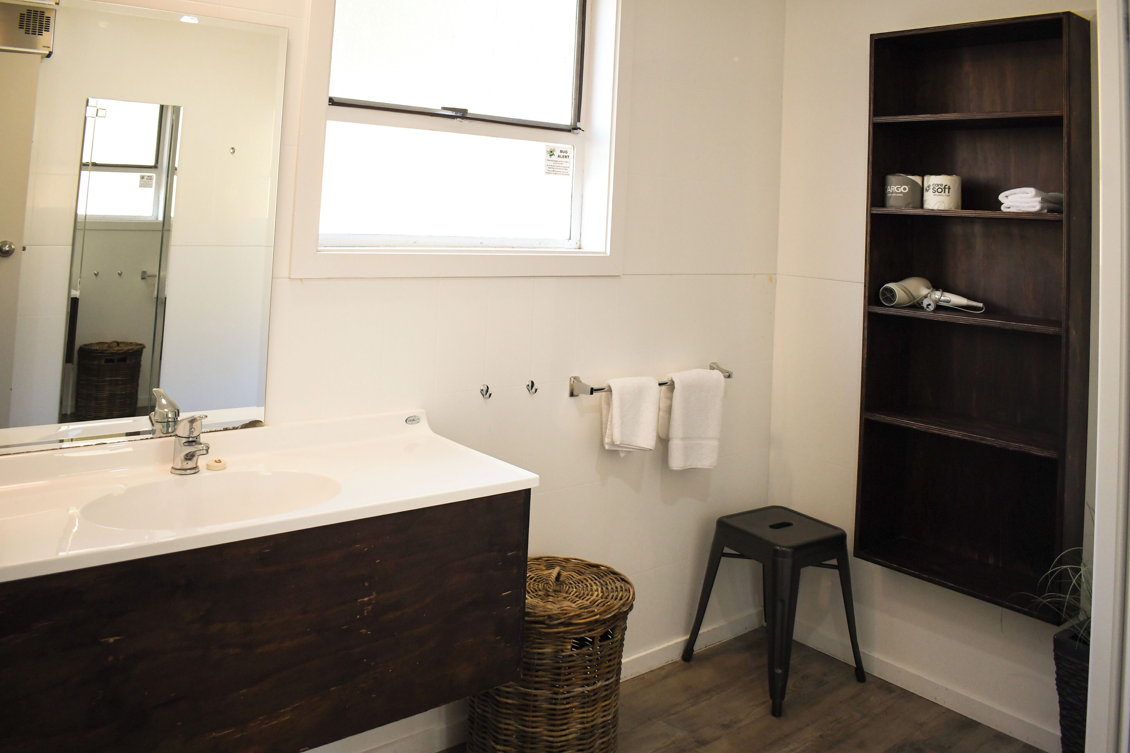 Un baño con un espejo grande, un lavabo blanco, un mueble de tocador de madera oscura, un taburete pequeño, una cesta de mimbre y una estantería con artículos de aseo.