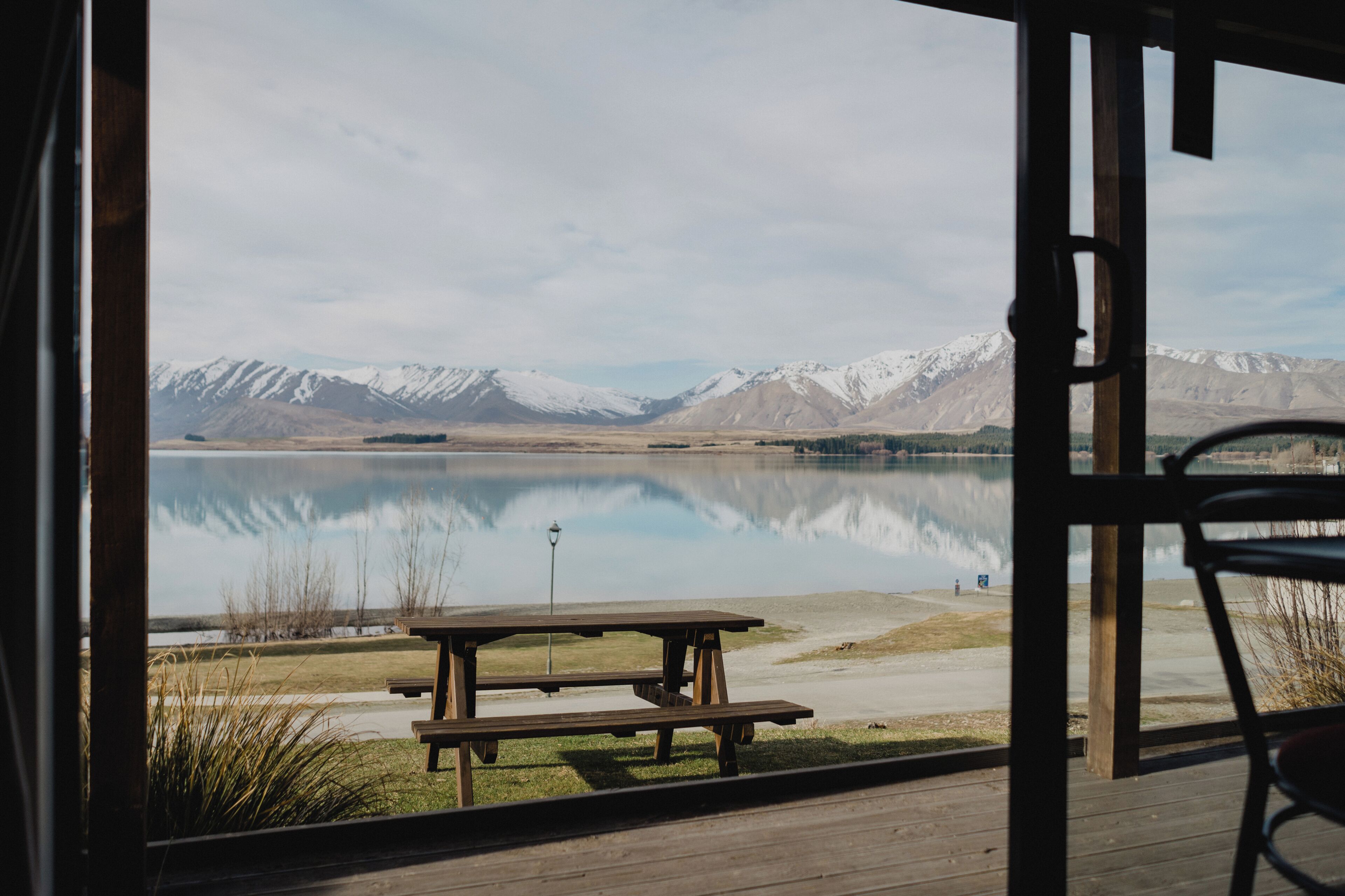 Une terrasse en bois avec une table de pique-nique et une chaise, donnant sur un lac paisible avec des montagnes en arrière-plan.