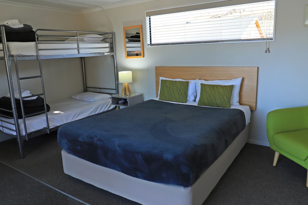 A hotel room with bunk beds, a single bed, a nightstand with a lamp, a green chair, and a window with blinds.
