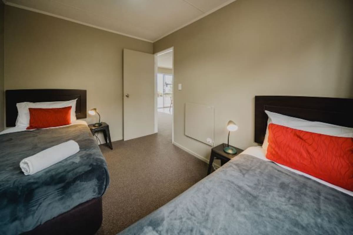 A hotel room with two beds, each with a different colored headboard and pillow. There is a small table with a lamp between the beds, and a door leading to another room.