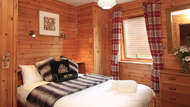 A wooden cabin bedroom with a bed, pillows, a nightstand, a window with curtains, a mirror, and a wall-mounted picture. - Private Harry Potter themed lodge with lovely views (Arisaig)