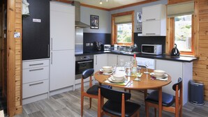 A compact kitchen with white cabinets, a microwave, and a stove. There is a dining table with chairs and a window with blinds. - Private Star Wars themed lodge with lovely views (Arisaig)