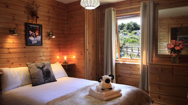 A wooden cabin bedroom with a bed, a nightstand with a lamp, a framed poster, a window with curtains, and a view of the outdoors. - Private Star Wars themed lodge with lovely views (Arisaig)