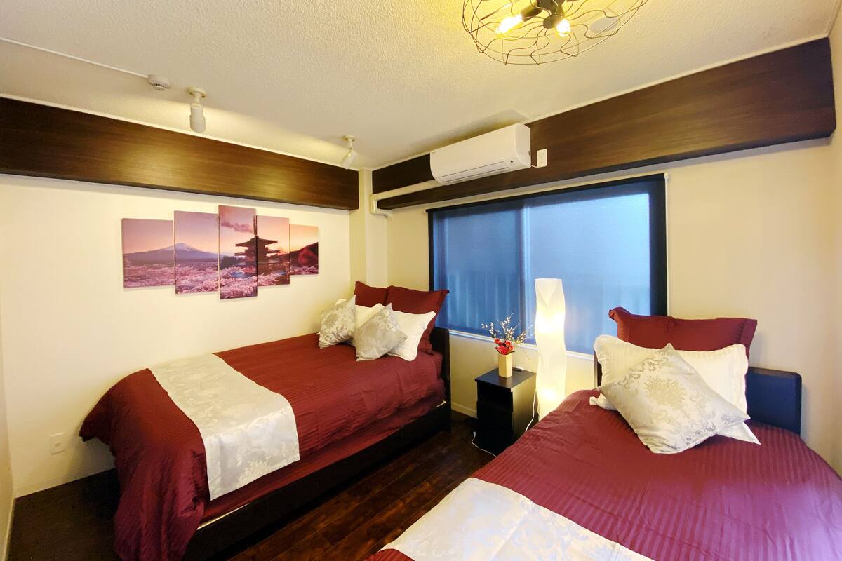 A hotel room with two beds, a large window with blinds, a ceiling fan, and wall-mounted air conditioning.