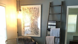 Un salon chaleureux avec un fauteuil en osier, un pouf en cuir noir, un tapis à motifs, une échelle et une estampe botanique encadrée accrochée au mur.