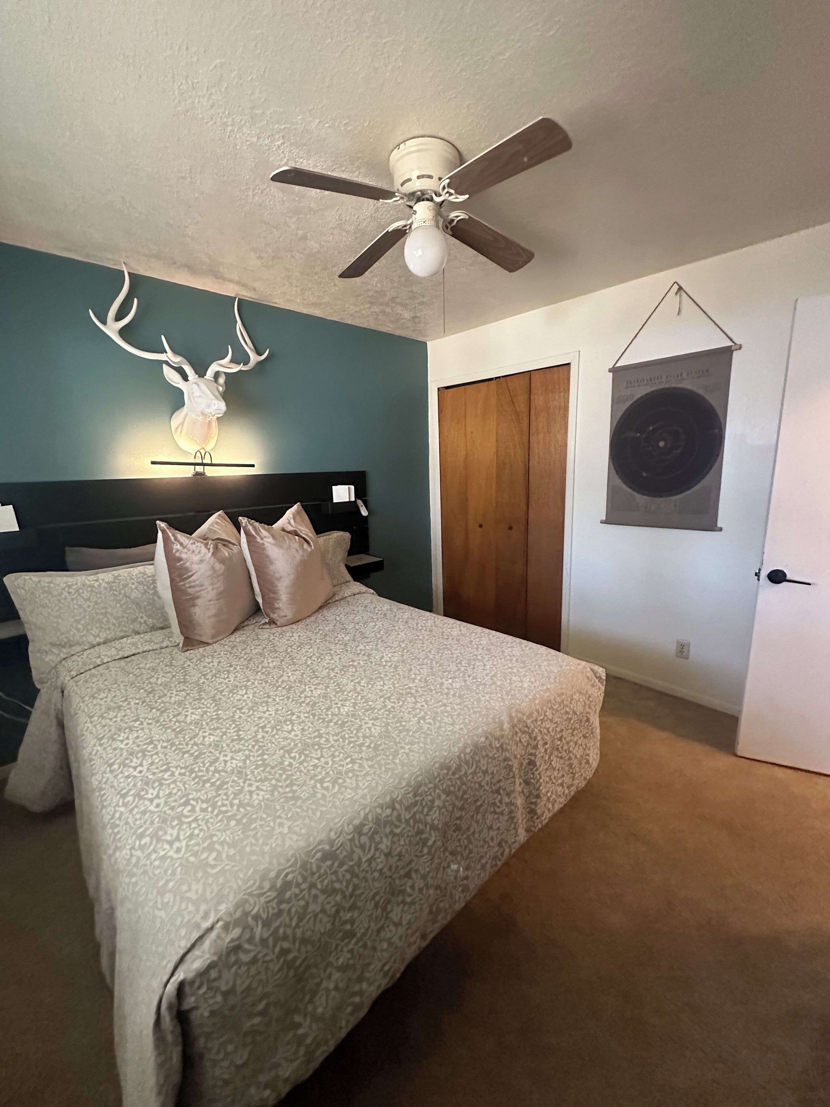 Een slaapkamer met een bed, een plafondventilator, een aan de muur bevestigd decoratiehoofd van een hert, een houten garderobe en een poster aan de muur.