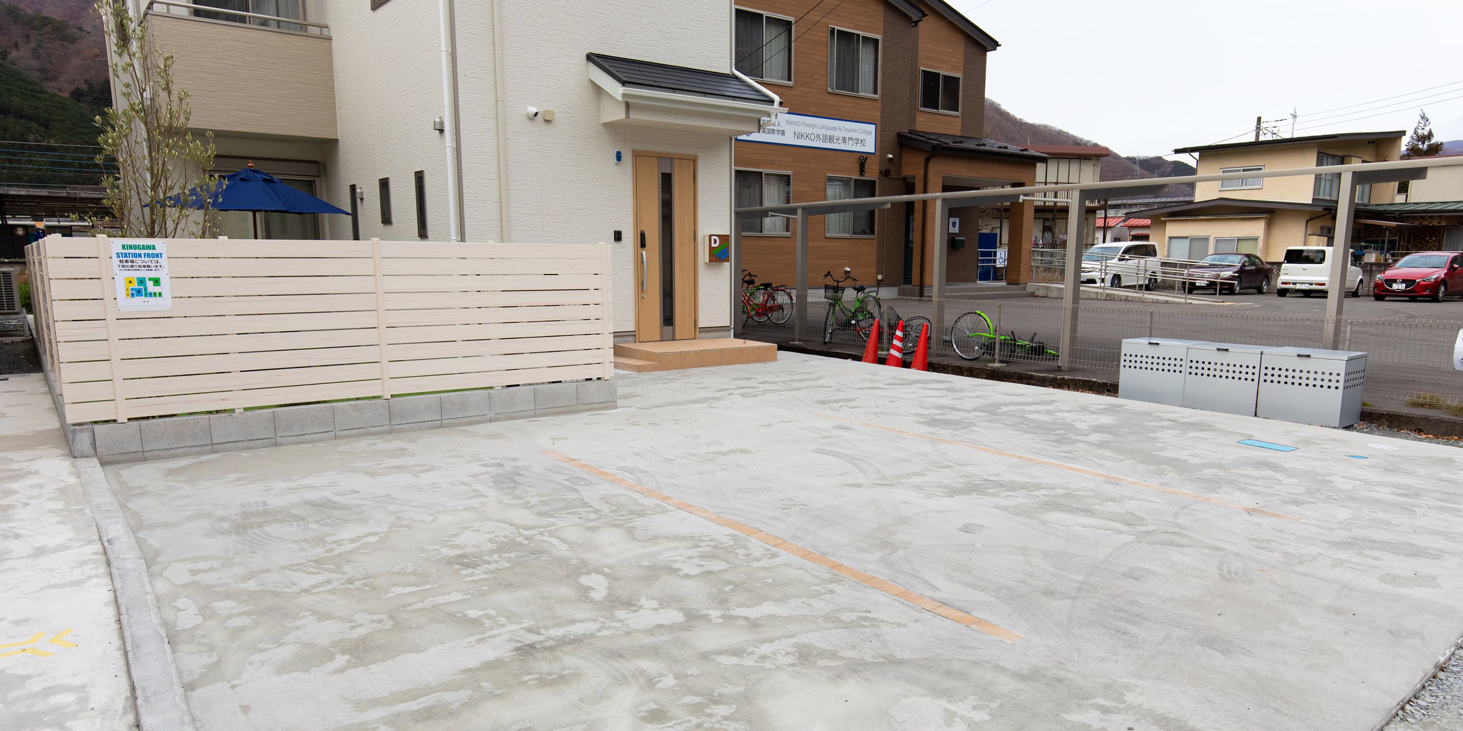 A parking area with a concrete surface, a wooden fence, and a building with a sign indicating it is the station front of Kinugawa.
