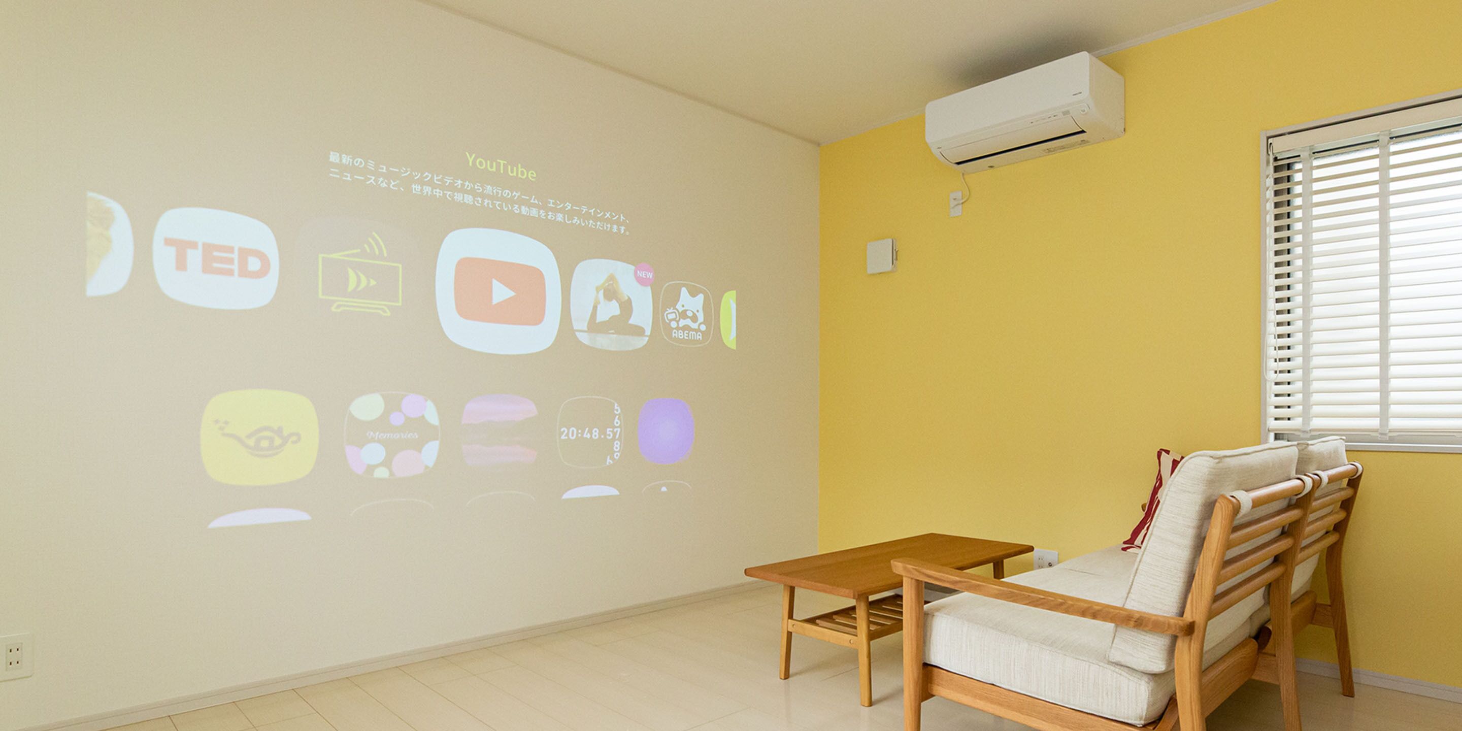 Quarto com parede amarela, uma cadeira de madeira, uma pequena mesa de madeira e uma tela de projeção exibindo diversos ícones.