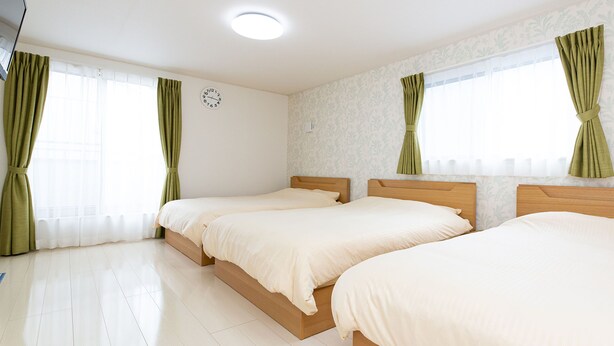A hotel room with two single beds, a wooden headboard, a white clock on the wall, and green curtains on the windows.