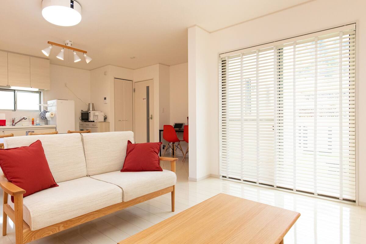 A modern kitchen with white cabinets, a wooden dining table, and a beige sofa with red cushions.
