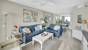 A spacious living room with a blue sofa set, a white coffee table, and a ceiling fan.
