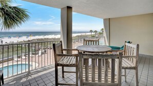 A balcony with a view of the beach, a pool, and wooden furniture.