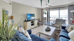 A spacious living room with a large TV, comfortable seating, a balcony with a view, and a ceiling fan.
