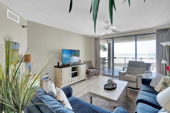 A spacious living room with a large TV, comfortable seating, a balcony with a view, and a ceiling fan.