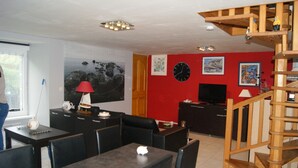 A dining area with a table and chairs, a red wall with a clock and paintings, a wooden staircase, and a TV on a cabinet. - Charming house set in grounds and forest, guaranteed peace and quiet. (Gommenec'h)