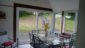 A dining area with a glass table and chairs, a vase with yellow flowers, and a view of a grassy area through large windows. - Charming house set in grounds and forest, guaranteed peace and quiet. (Gommenec'h)